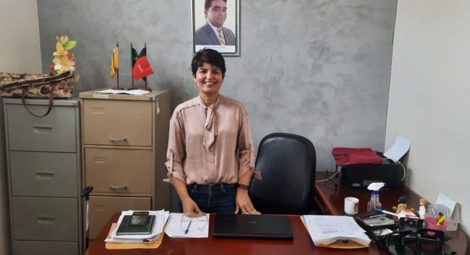 Emanuelle, Ex Secretária de Saúde de Mari, em uma sala, expressando felicidade com uma foto do Prefeito AG na parede.