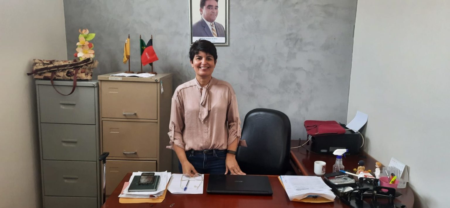 Emanuelle, Ex Secretária de Saúde de Mari, em uma sala, expressando felicidade com uma foto do Prefeito AG na parede.
